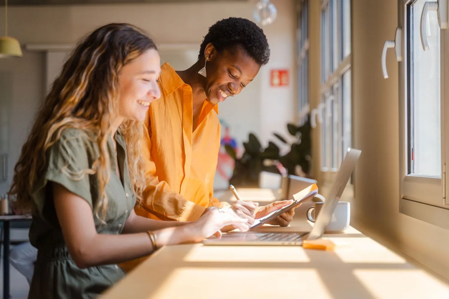 Two colleagues smiling and reviewing work together at a sunny desk, one taking notes while the other points at a laptop in a bright modern office.