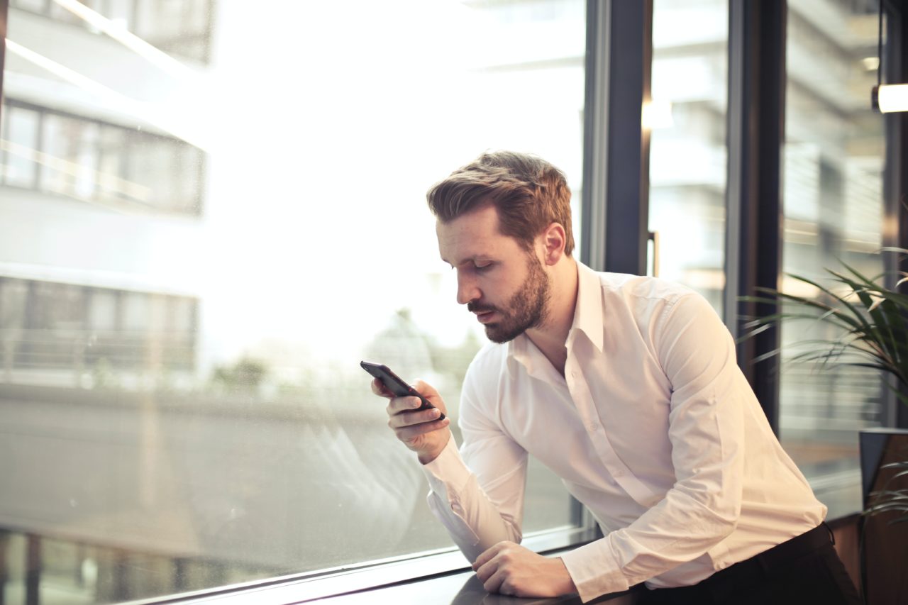 Man in office leans on window and browses custom intranet on smartphone.