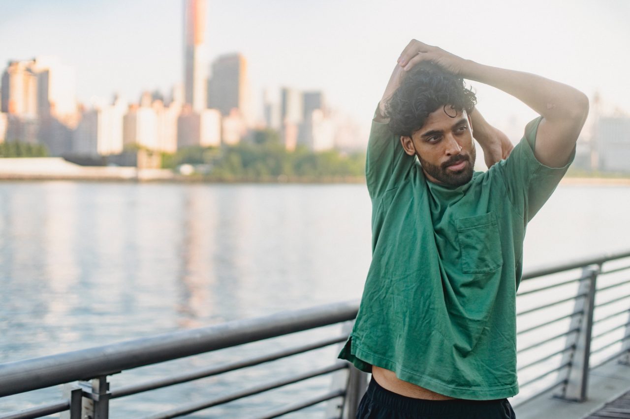 A male jogger stretches his arms by a river with a city skyline in the background.