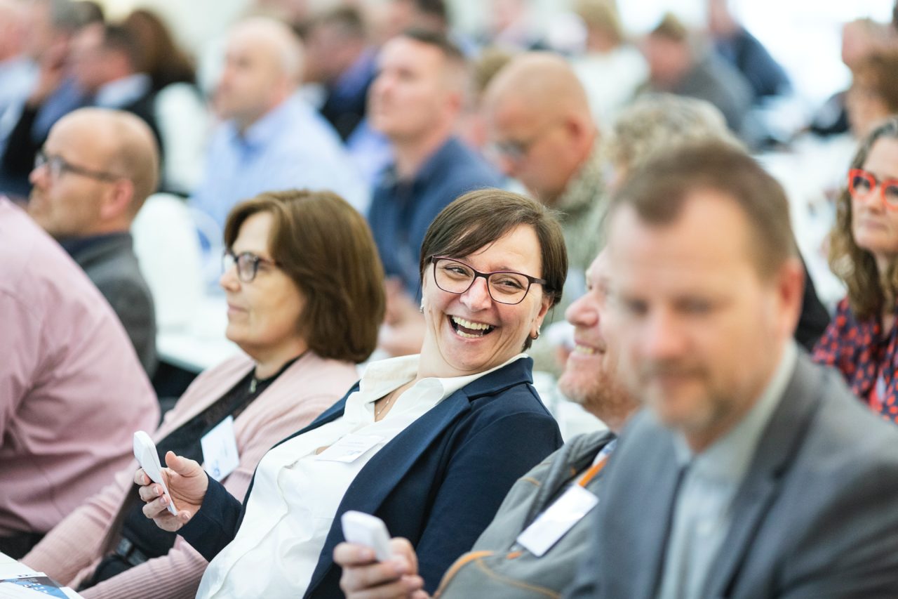 A middle aged woman and man laugh together sitting among the crowd at a corporate event.