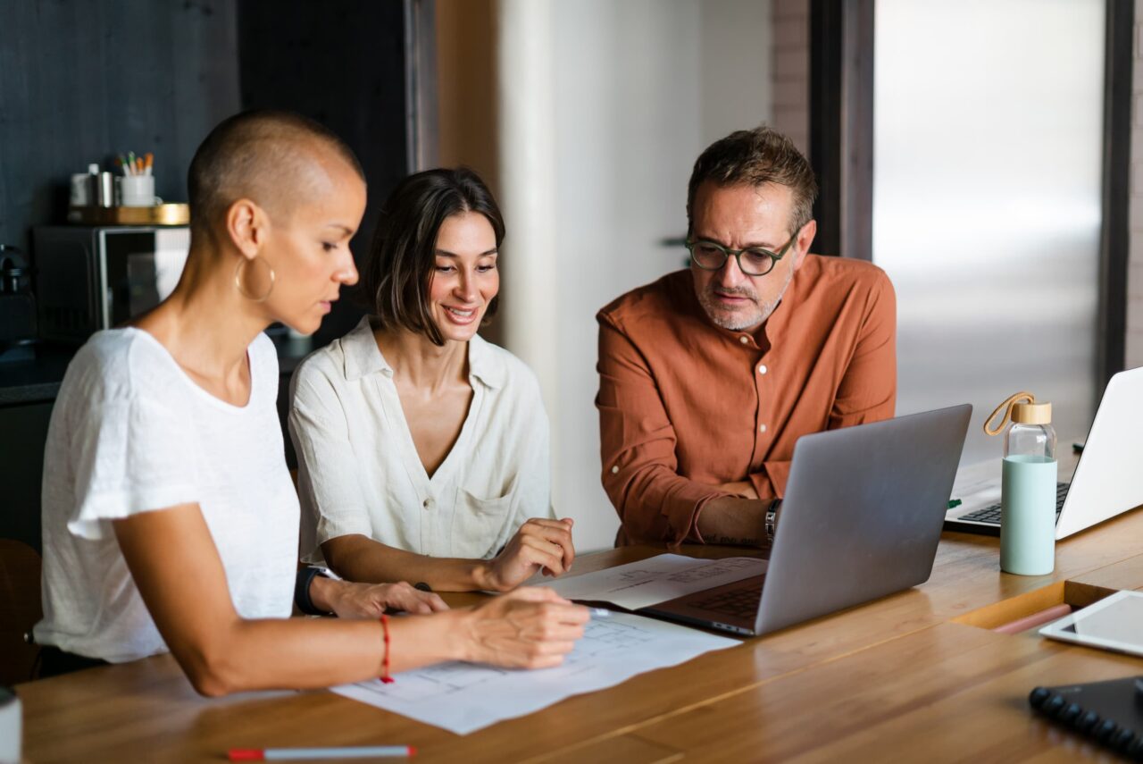 Three colleagues reviewing intranet professional services plans together around a laptop and printed documents at a wooden table in a modern workspace.