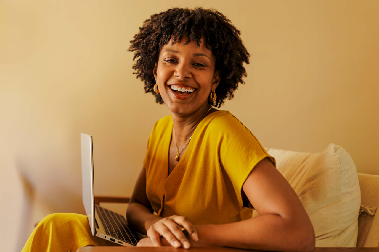 A woman in a yellow dress laughing warmly while working on a laptop in a warm home office setting, conveying a positive and engaged remote work experience.