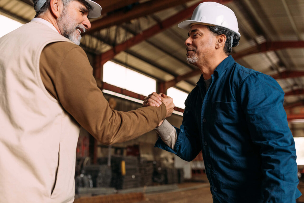 Two workers in hard hats shaking hands inside an industrial warehouse, representing the trusted professional services partnership Interact builds with customers across sectors.