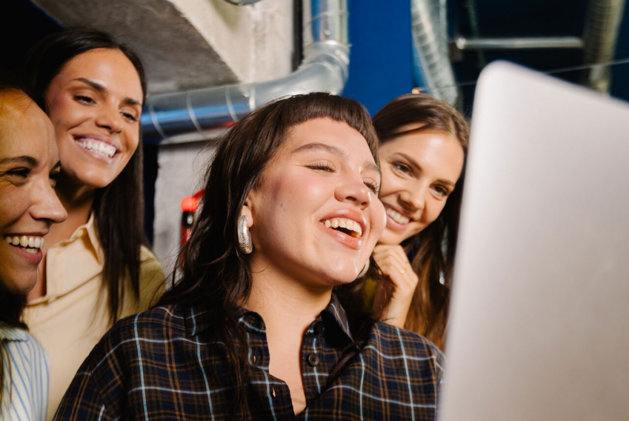 Four women laughing together while looking at a laptop screen in an industrial-style office, conveying a positive and energetic team culture.