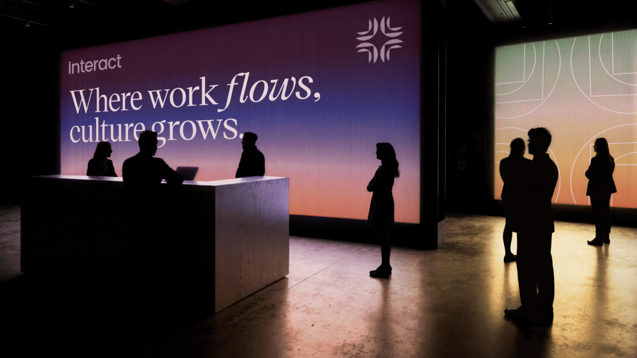 The employee experience journey brought to life at an Interact event, with silhouetted attendees gathered beneath a large screen displaying "Where work flows, culture grows."
