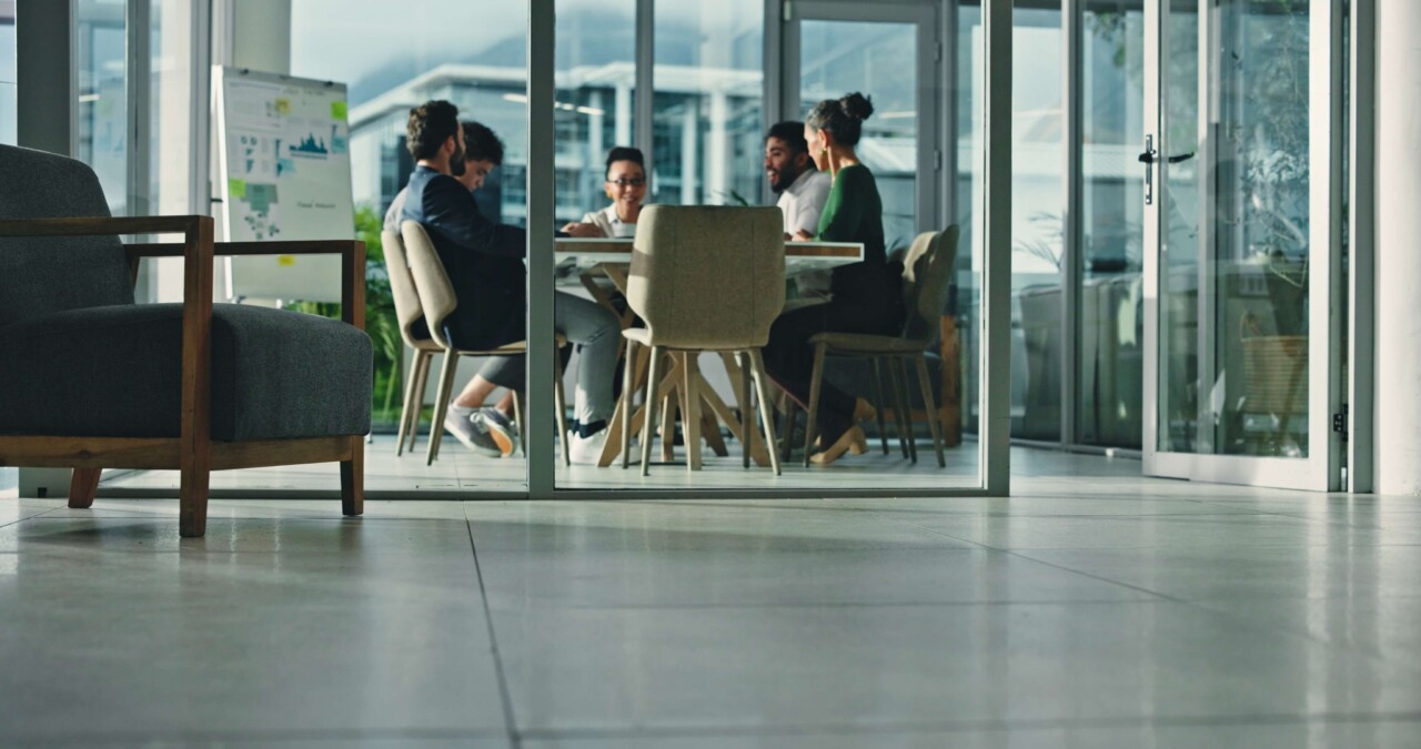 An inclusive workplace in action, with four colleagues collaborating around a table in a bright glass-walled meeting room with a data-covered whiteboard.