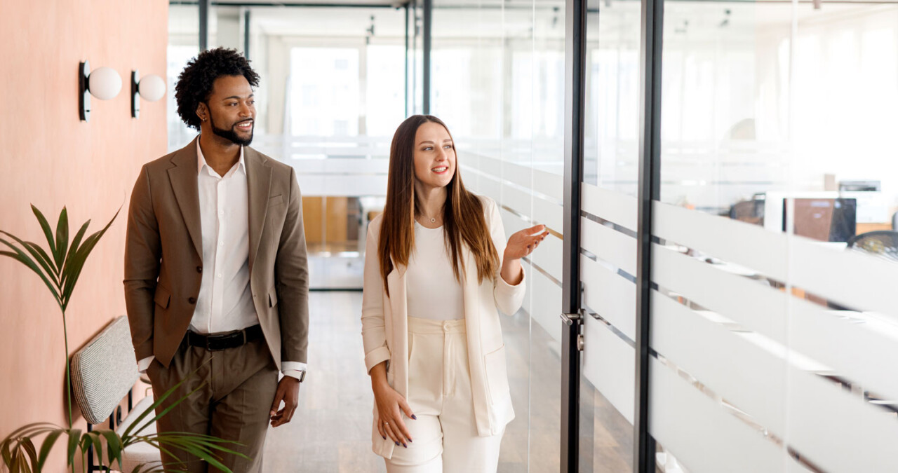 Two colleagues walking and talking in a bright modern office corridor, conveying an open and collaborative working environment.