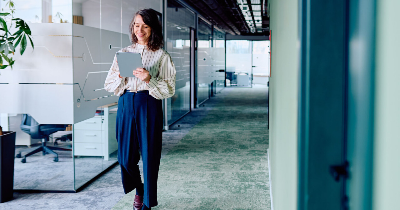 A woman in a striped blouse smiling at a tablet while leaning against a glass partition in a modern open-plan office hallway.