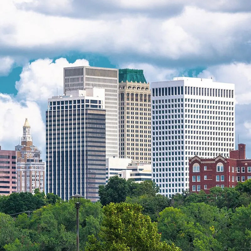 Tulsa city skyline viewed from across a tree line, with a mix of modern glass towers and a historic terracotta-roofed skyscraper under a partly cloudy sky.