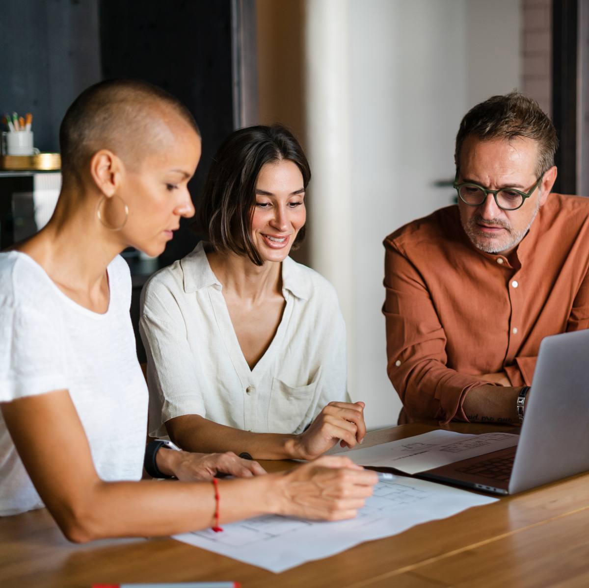 Three colleagues reviewing intranet strategy services plans together around printed documents and a laptop at a wooden table in a relaxed modern workspace.