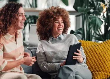 Two colleagues laughing together on a sofa while reviewing content on a tablet, surrounded by indoor plants in a relaxed and informal workspace.