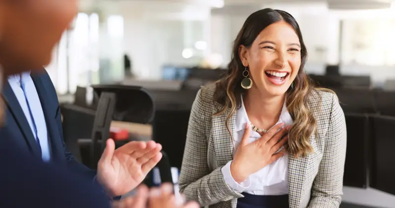 A woman in a blazer laughing warmly during a conversation in an open-plan office, conveying an energetic and people-first workplace culture.