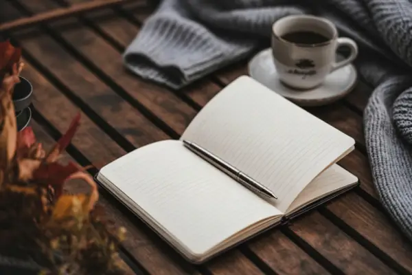 An open blank notebook with a pen resting on a rustic wooden table, alongside a coffee cup and a gray knit throw, evoking a calm and reflective working environment.