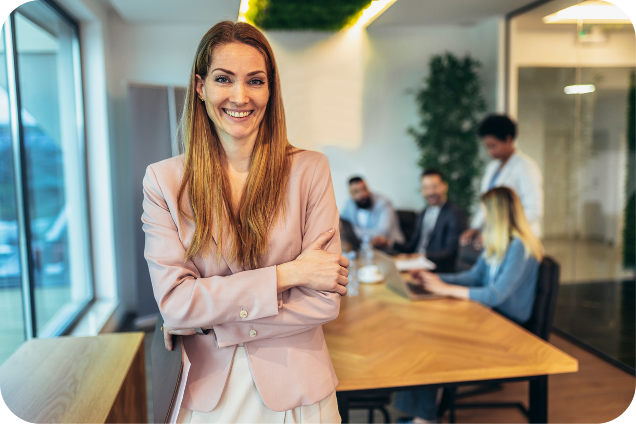 A smiling woman in a pink blazer standing with arms crossed in front of a team meeting in a glass-walled boardroom, conveying confident leadership.