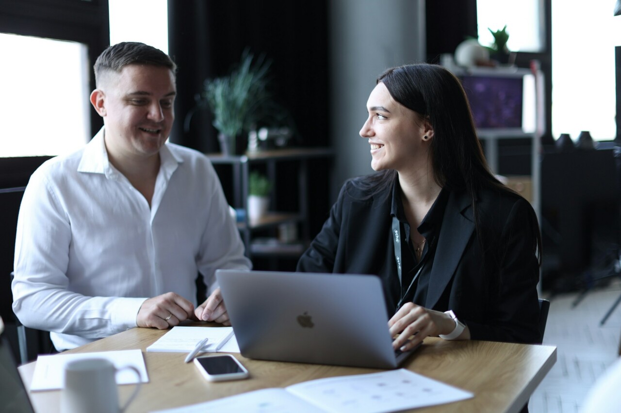 Two people sit at a computer in an office, with one asking another a question, representing an intranet software demo. 